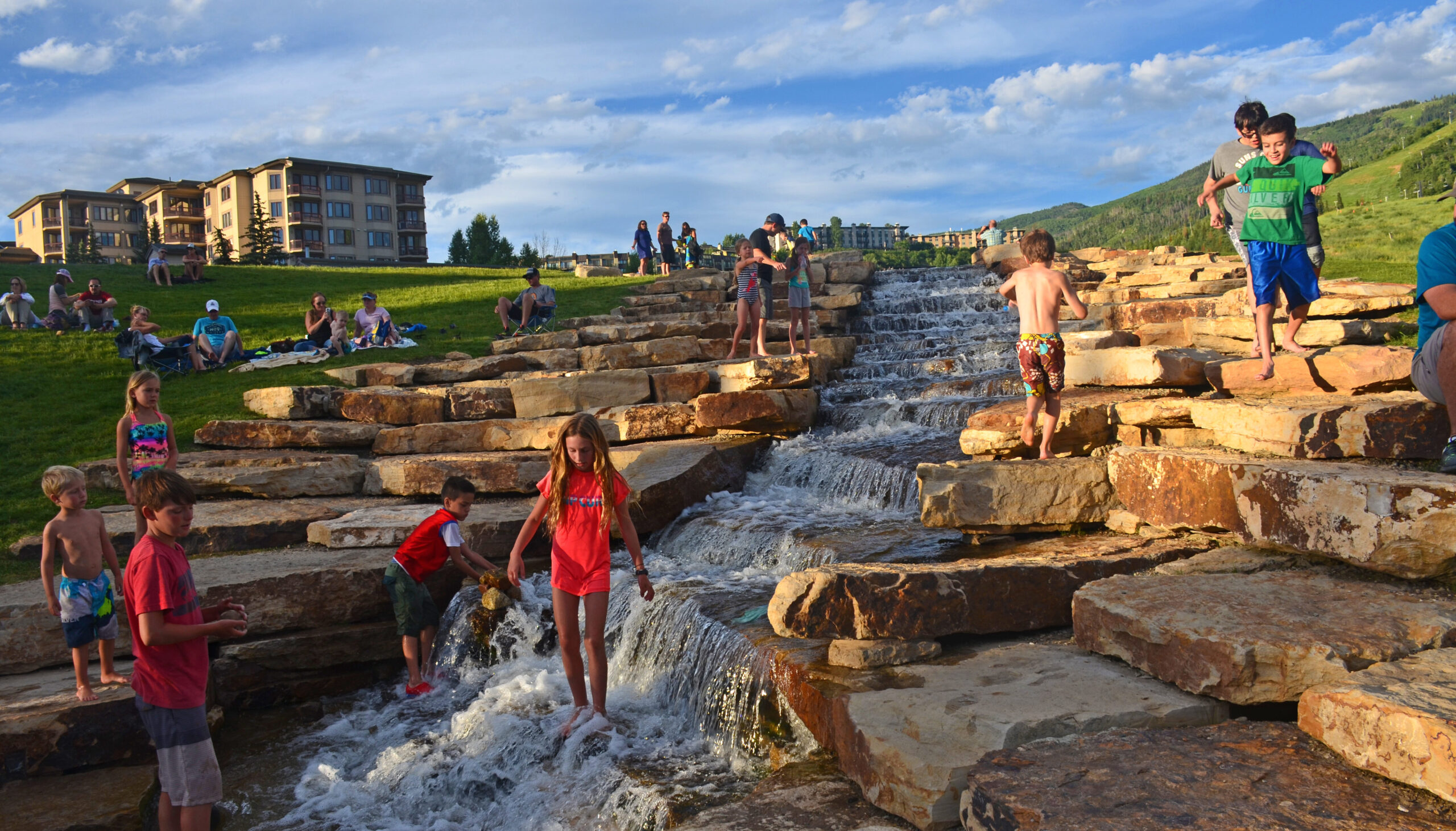 Children playing on stone waterfall steps at a scenic resort environment, people relaxing on grass in the background