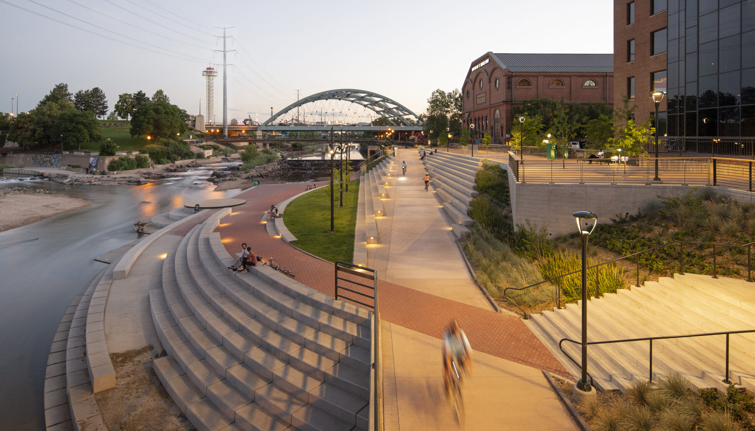 Confluence Park with cyclists and pedestrians, evening ambiance, Shoemaker Plaza steps, distant bridge and trees.