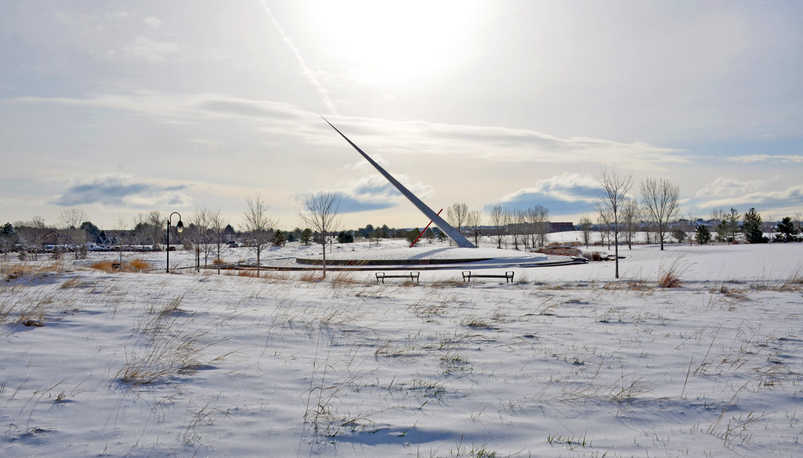 Snow-covered park with large sundial sculpture and bare trees under a cloudy sky