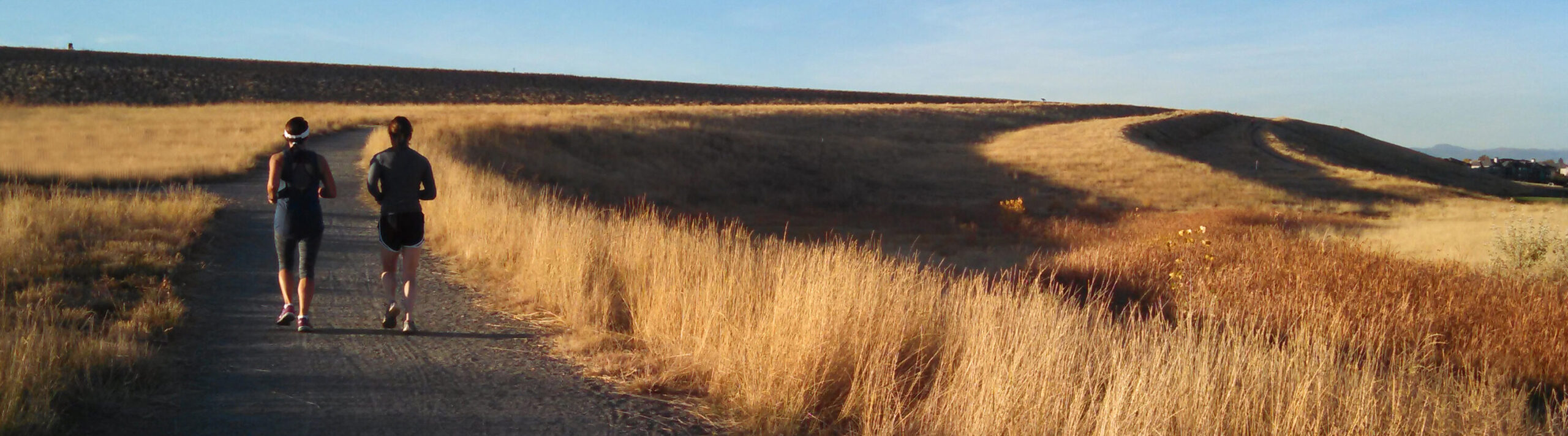 Two people jogging on a path through grassy hills under a clear sky
