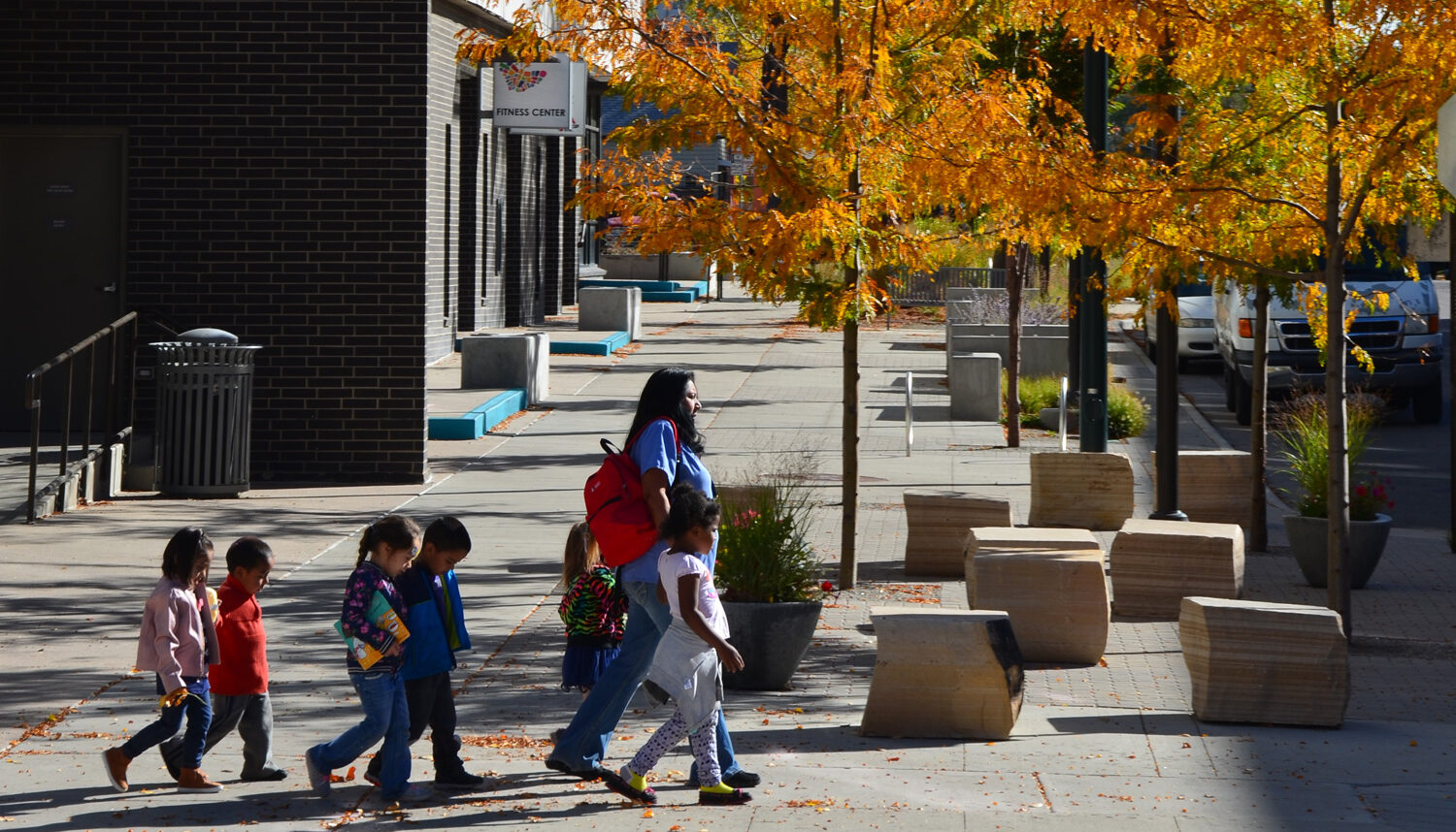 A line of young children walk by an assortment of cubic sandstone seats and orange honeylocust trees.