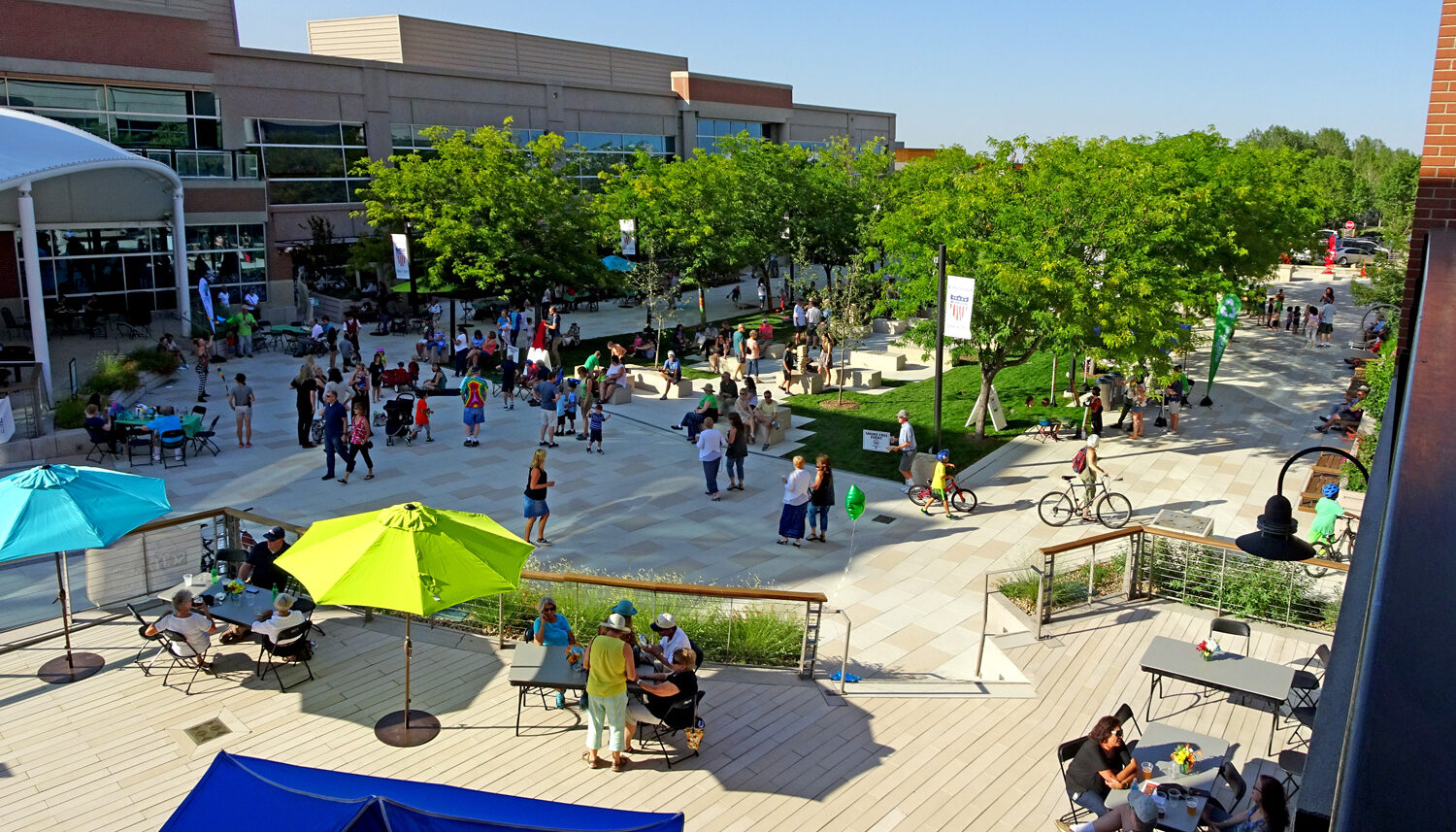 A photo shows a plaza with a large turf lawn in the center surrounded by trees and with stone paving and stone seatwalls within it. People are milling, walking, and dining.