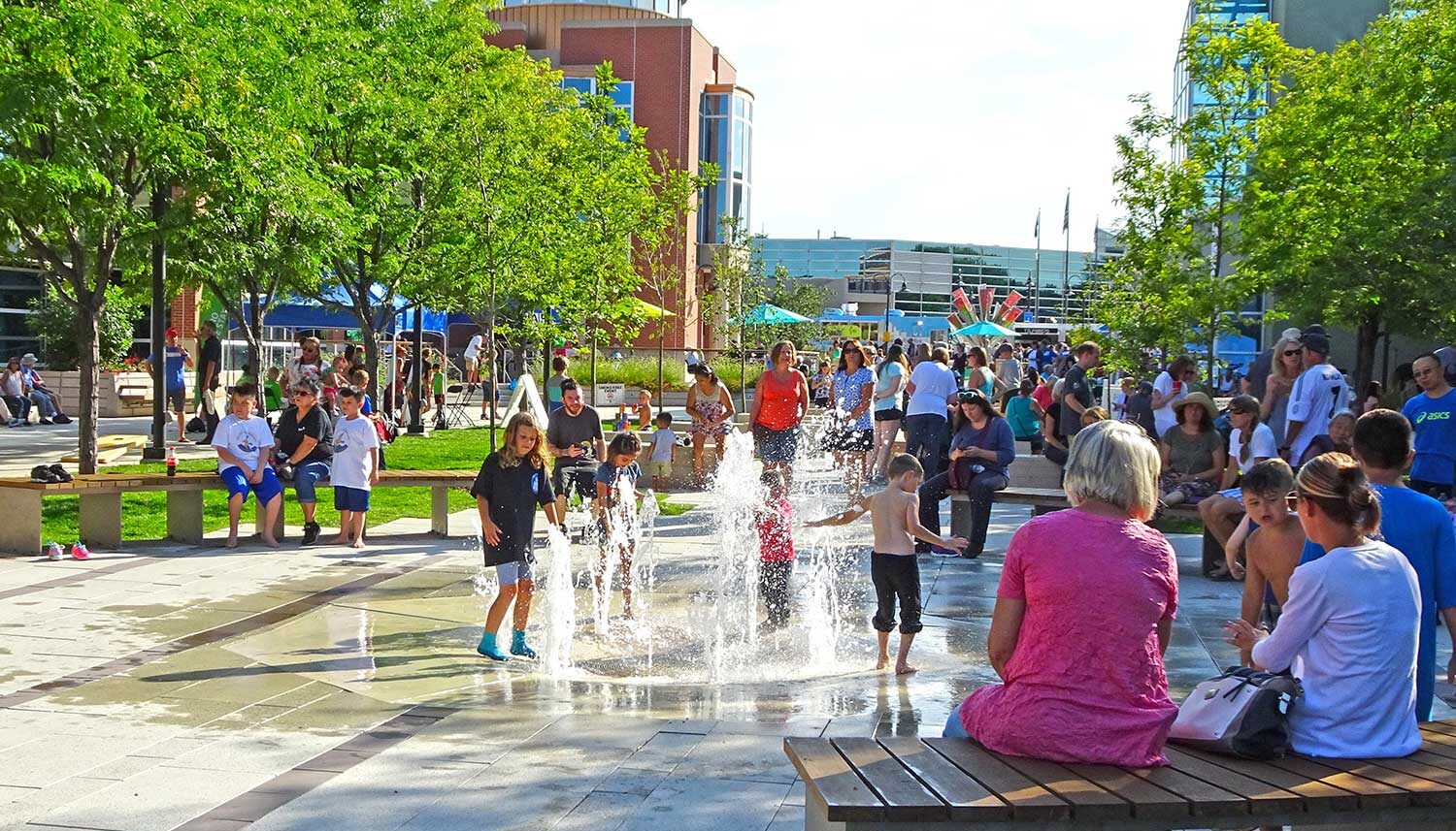 Stone paving with jets of water shooting form the ground entertain a small group of kids while adults watch from surrounding seats.
