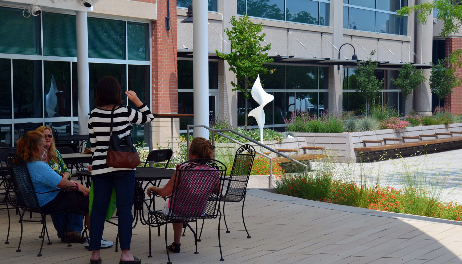 A group of women sit at a cafe table in the shade with a series of raised planters and the plaza behind.