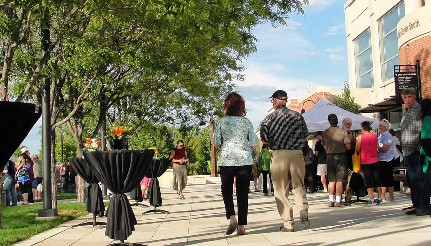 People mill between event tables and tents beneath mature honeylocust trees.,