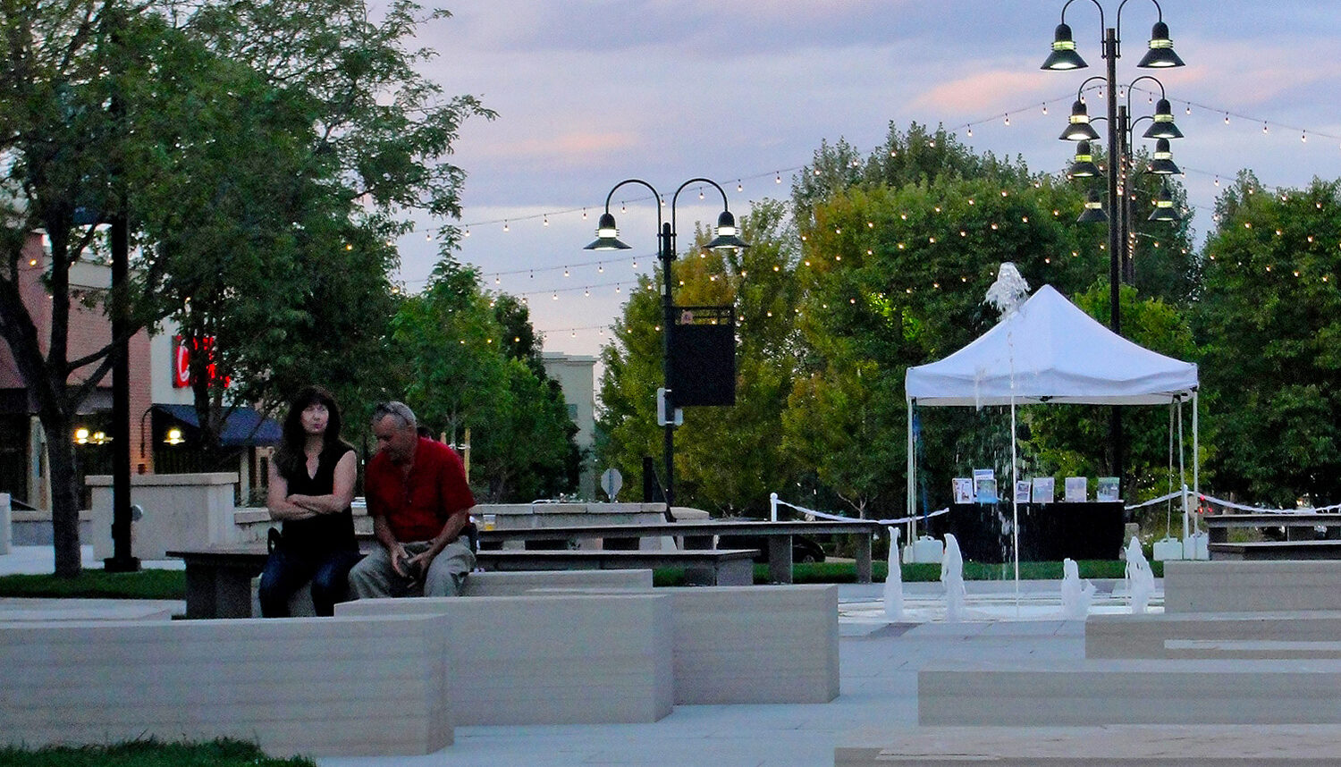 In dusk light, a couple sit on stone seats with hanging lights, tents, trees, and water jets behind them.