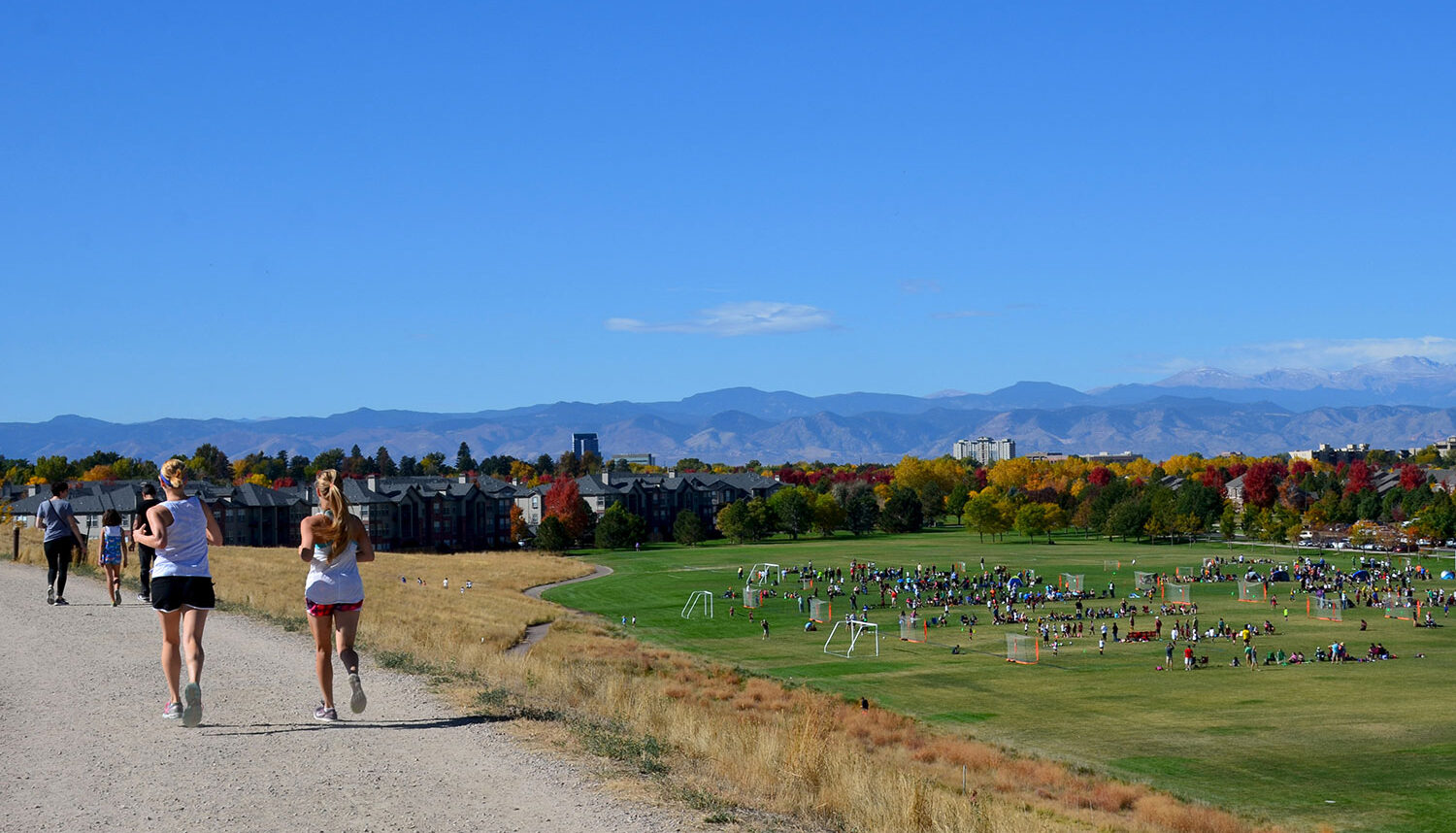 Runners on a gravel path run away from the photographer with soccer fields behind and the Rocky Mountains in the background.