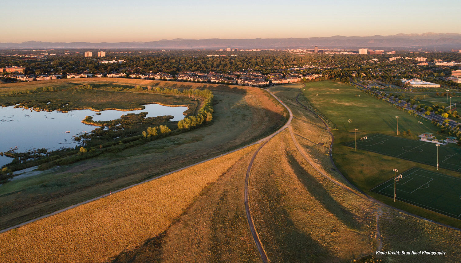 A drone photograph of the raised berm with gravel paths on top stretches into the distance toward the Rocky Mountains.