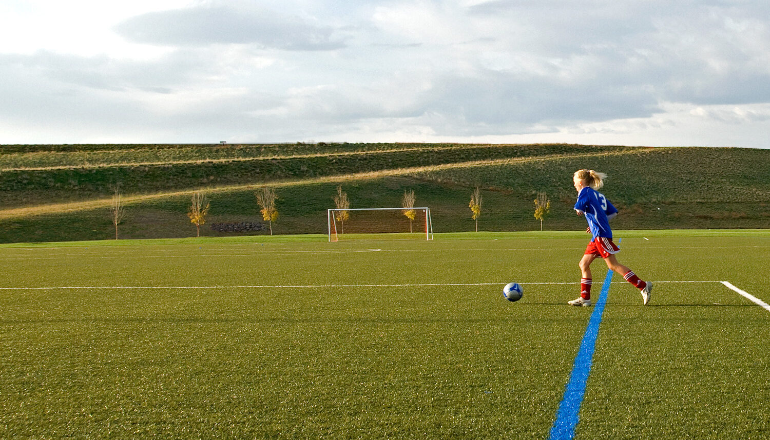 A young soccer player dribbles on the turf with young trees, a soccer goal, and the berm in the background.