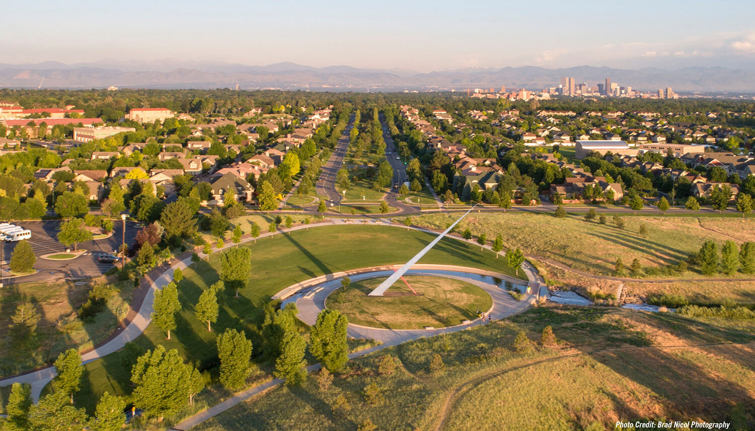 An aerial photo shows walking paths in organic shapes conform to a circular dias of turf which houses a large monument shaped like a needle with Denver in the background.
