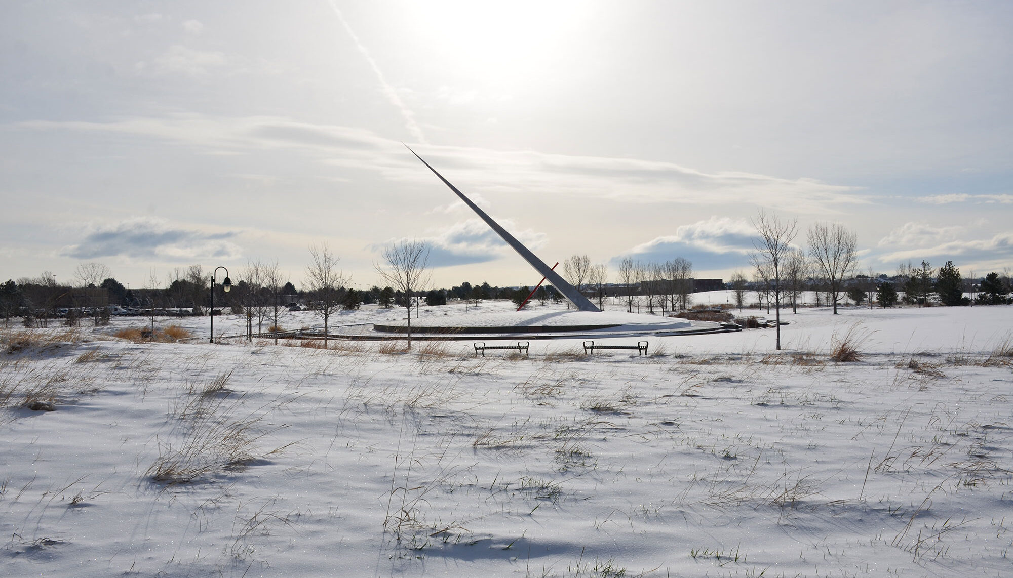 A photo of a winter field with a large dias in the background from which a massive sculpture points the the sky like a giant needle.