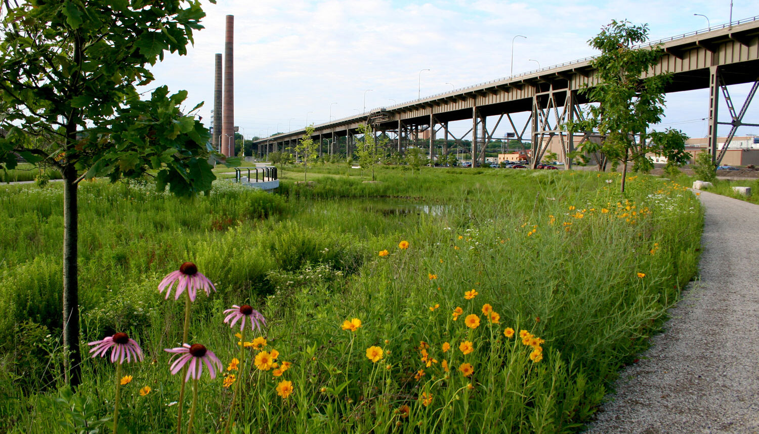 A photo shows purple and yellow flowers in the foreground of a lush, green field behind which is an overlook to observe a small pond. In the background is a pair of smokestacks and a raised roadway up on struts and trusses.