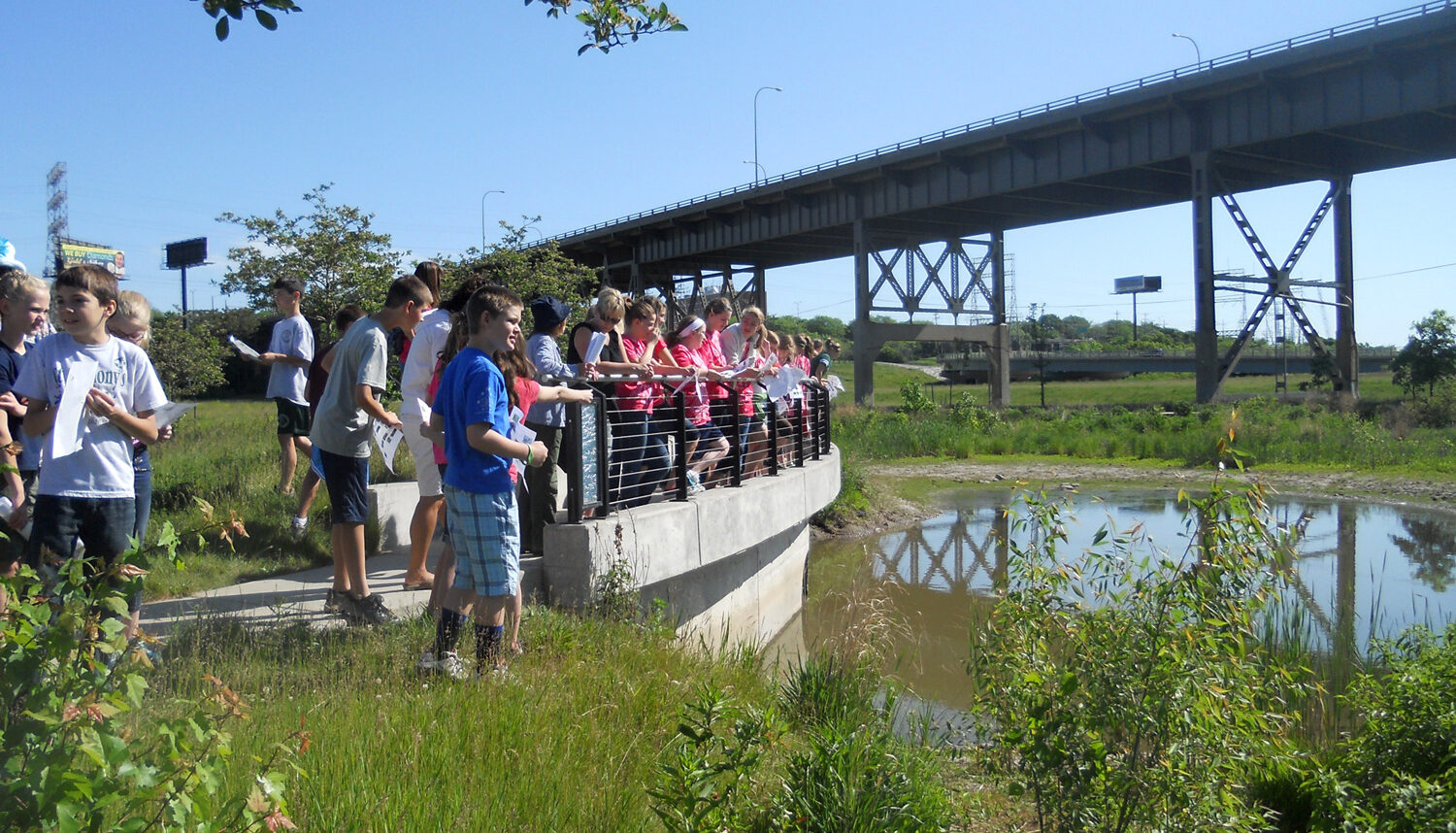 Children line up on a concrete overlook to view a small pond with the raised highway in the background.