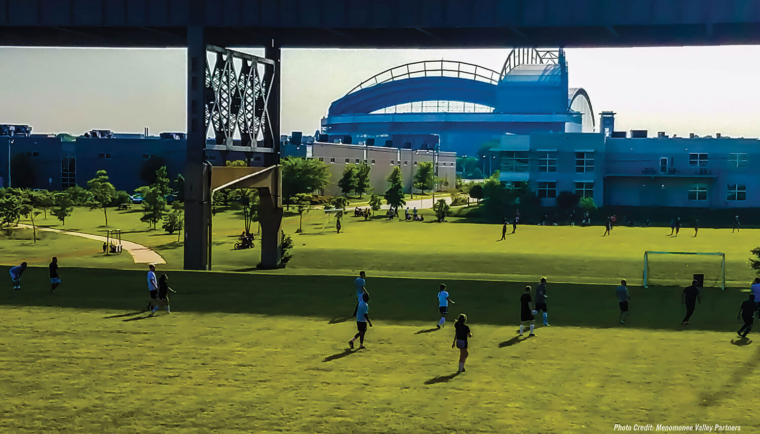 People play soccer on a large field cut in half by the shadow of the raised highway.