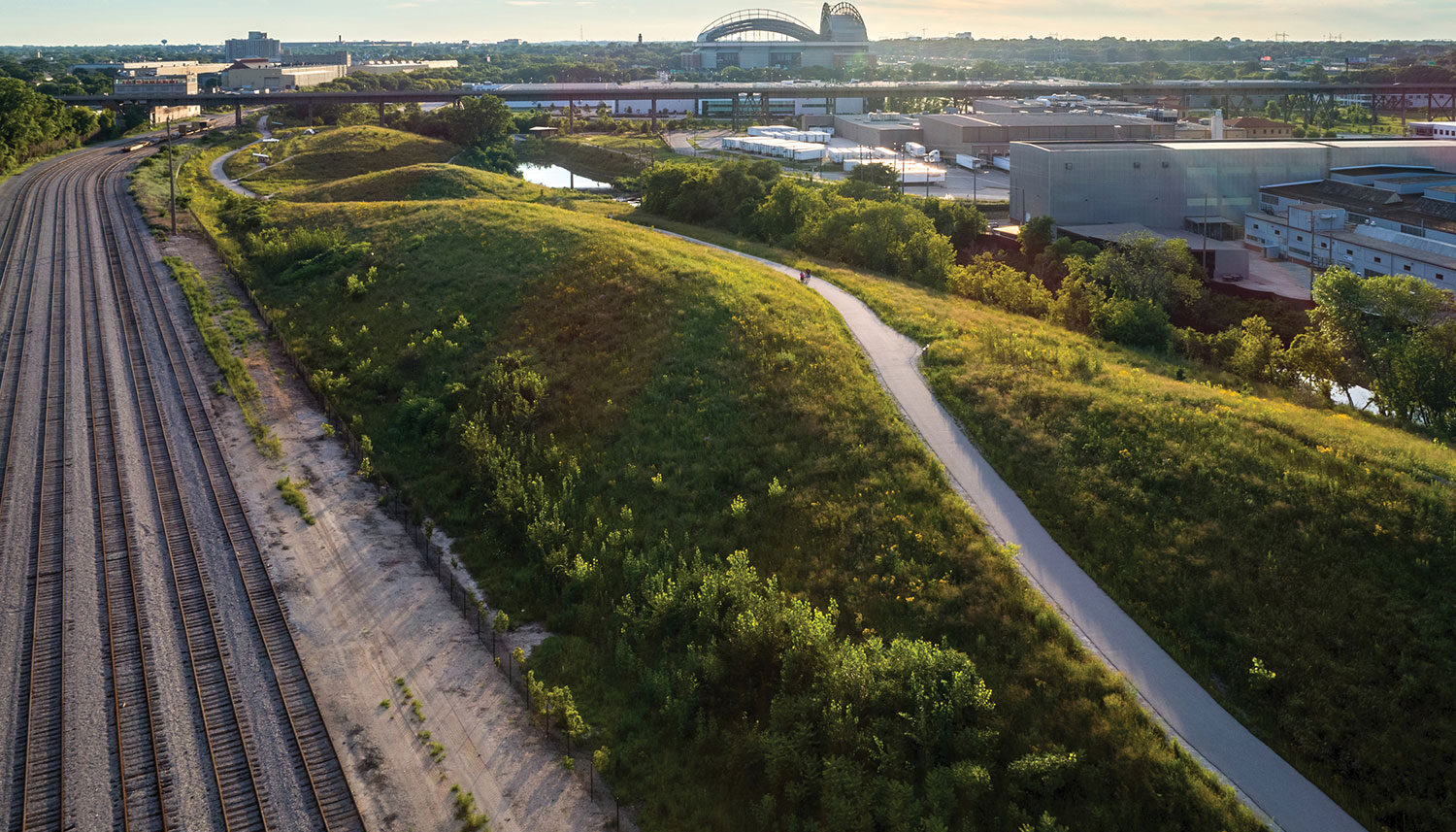 An aerial photo has rows of train tracks on one side of the image with rolling hills of green fields and a pedestrian path on the other side.