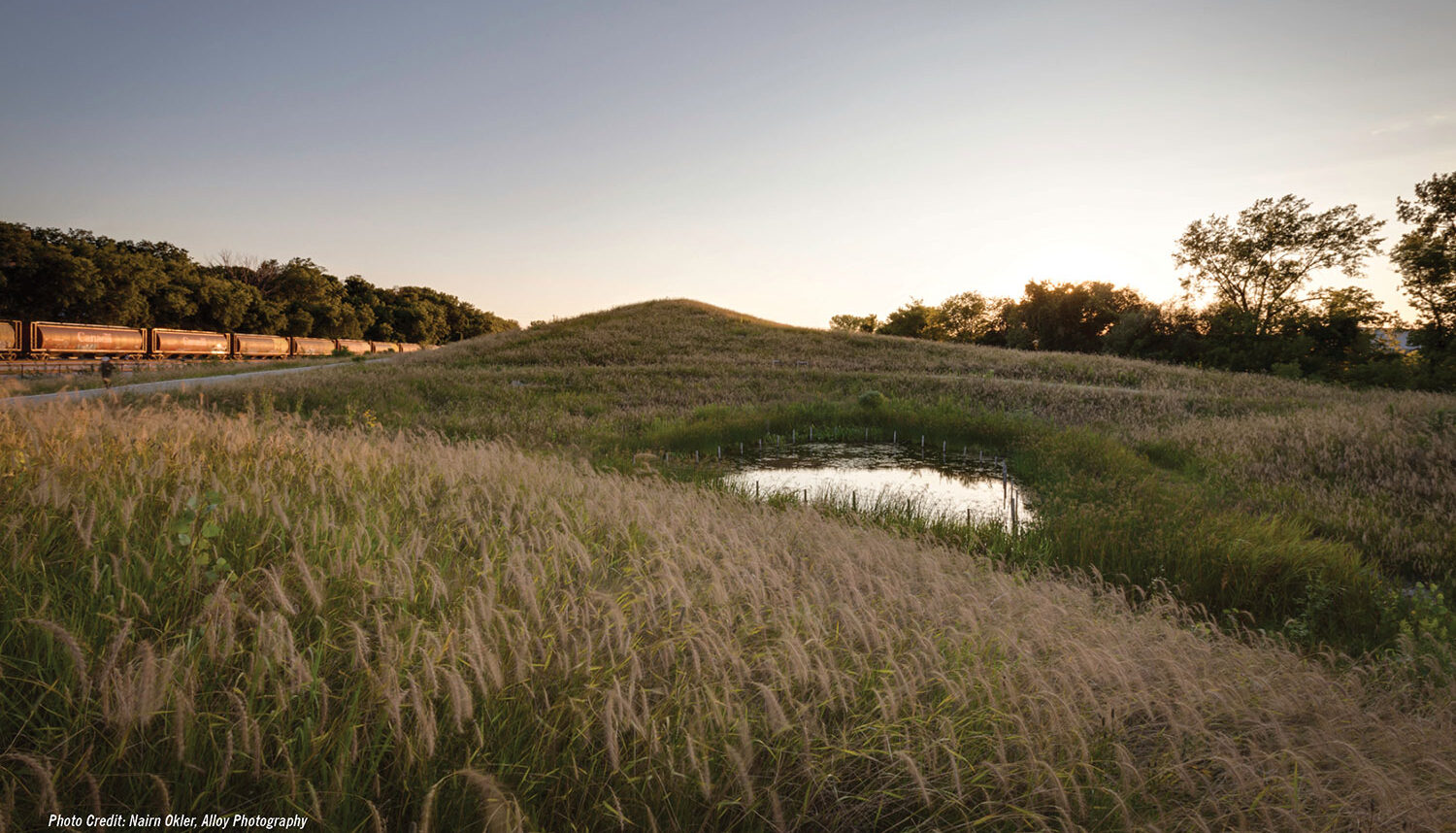In dusk light, a photo captures rolling hills of airy grasses nestling a small pond into its low point with a series of train cars resting in the background.