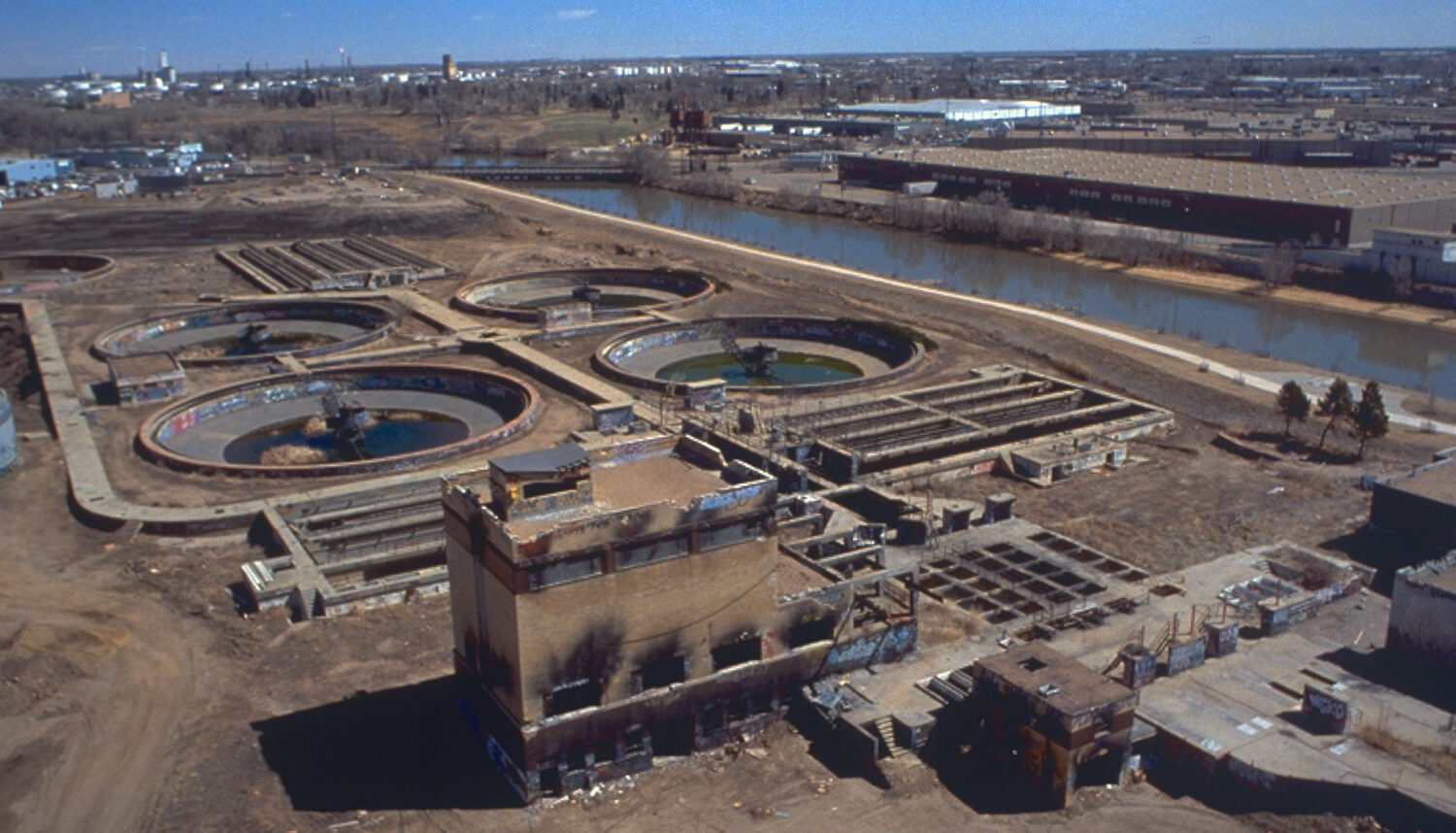An aerial photo shows the site before improvements: an industrial yard with four large concrete wells beside the river.
