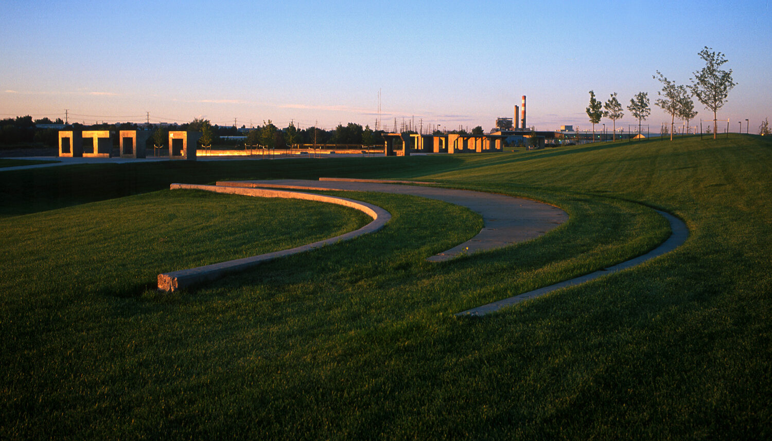 Green turf grass is broken by three semi-circular concrete bands with large, rectangular, concrete forms catching sunset light in the background.