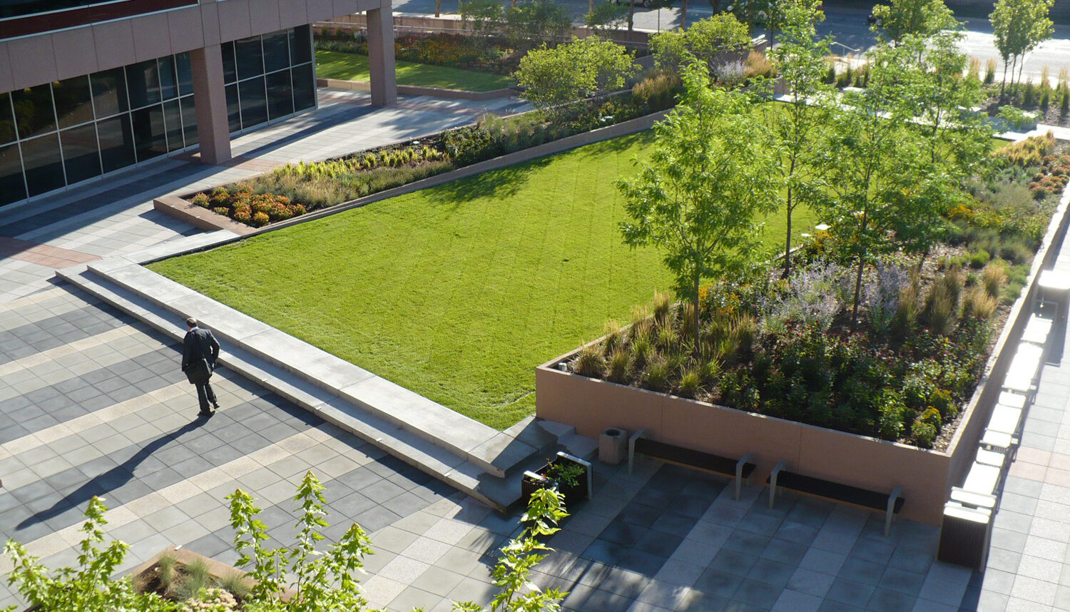 Framed by grey, concrete unit pavers, an elevated rectangle of turf grass is edged with raised planting beds.