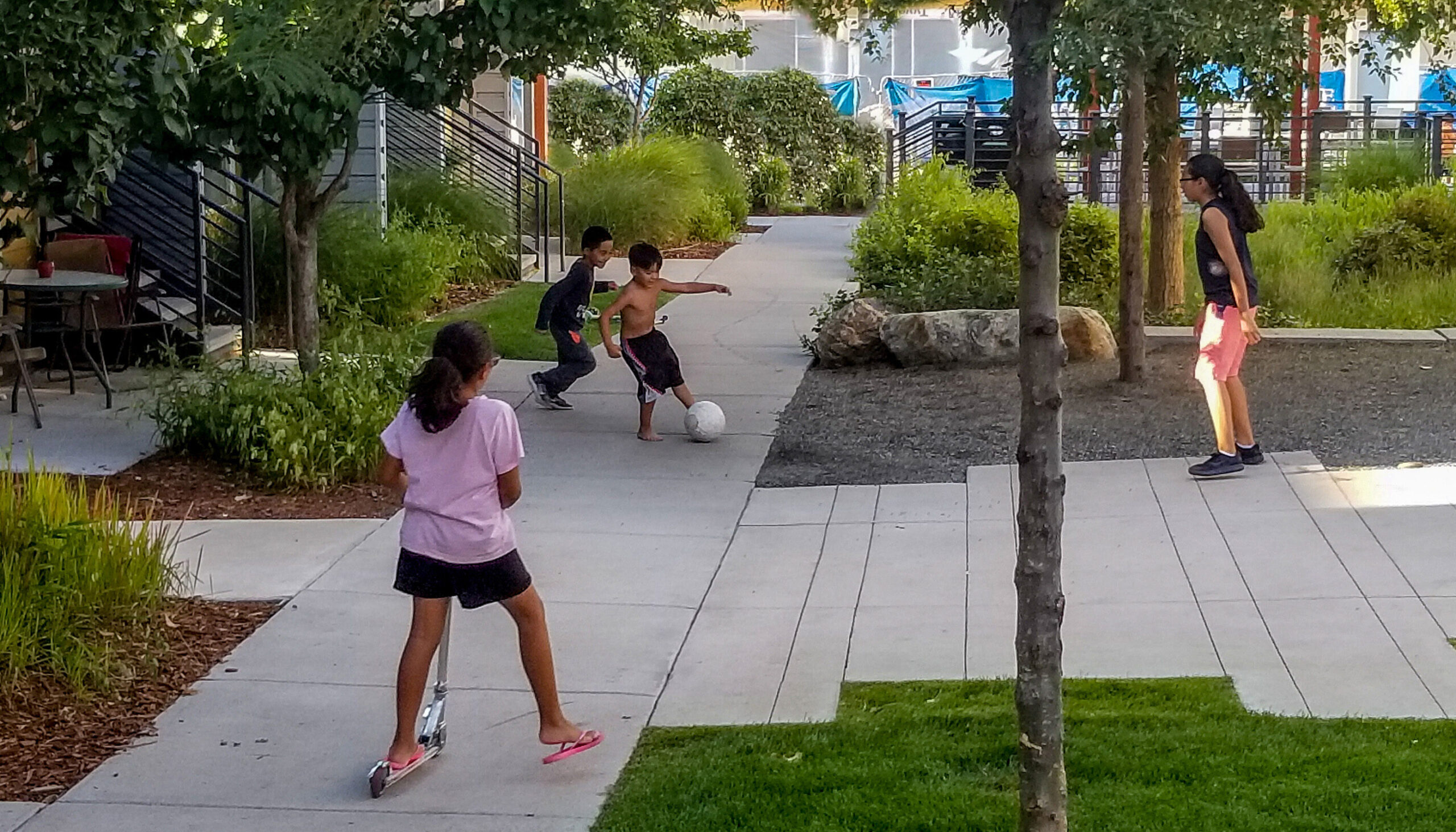kids playing soccer and riding a scooter in a shaded courtyard open space