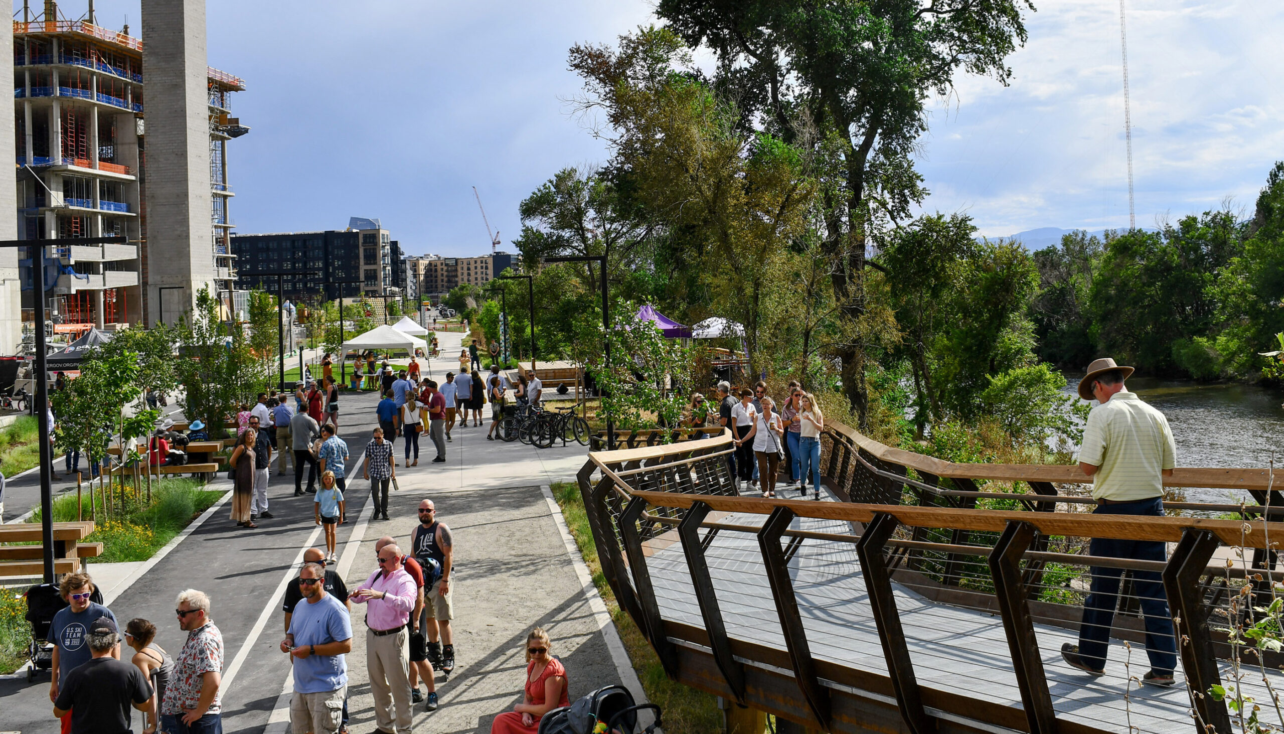 A view from the elevated walkway during a community event highlights on the left the many spaces people can gather along the linear park - i.e picnic tables, under tents, and on benches surrounded by plantings - and on the right, the South Platte River.