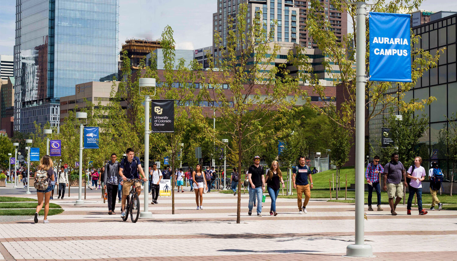 Students walk and bike along a banded plaza with campus flags and trees in the foreground and Denver in the background.