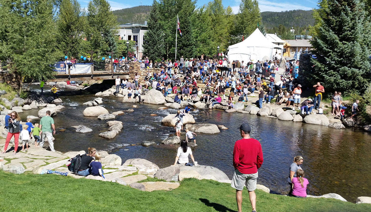 A stone-lined river is crowded with people attending and event with tents in the background.
