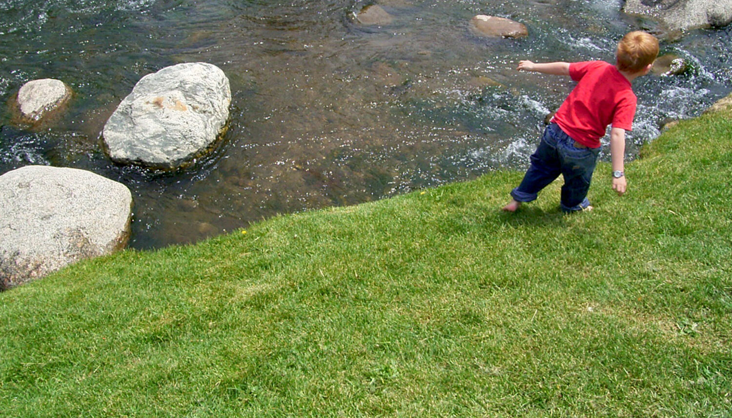 A young child enjoys the grass barefoot near the river.