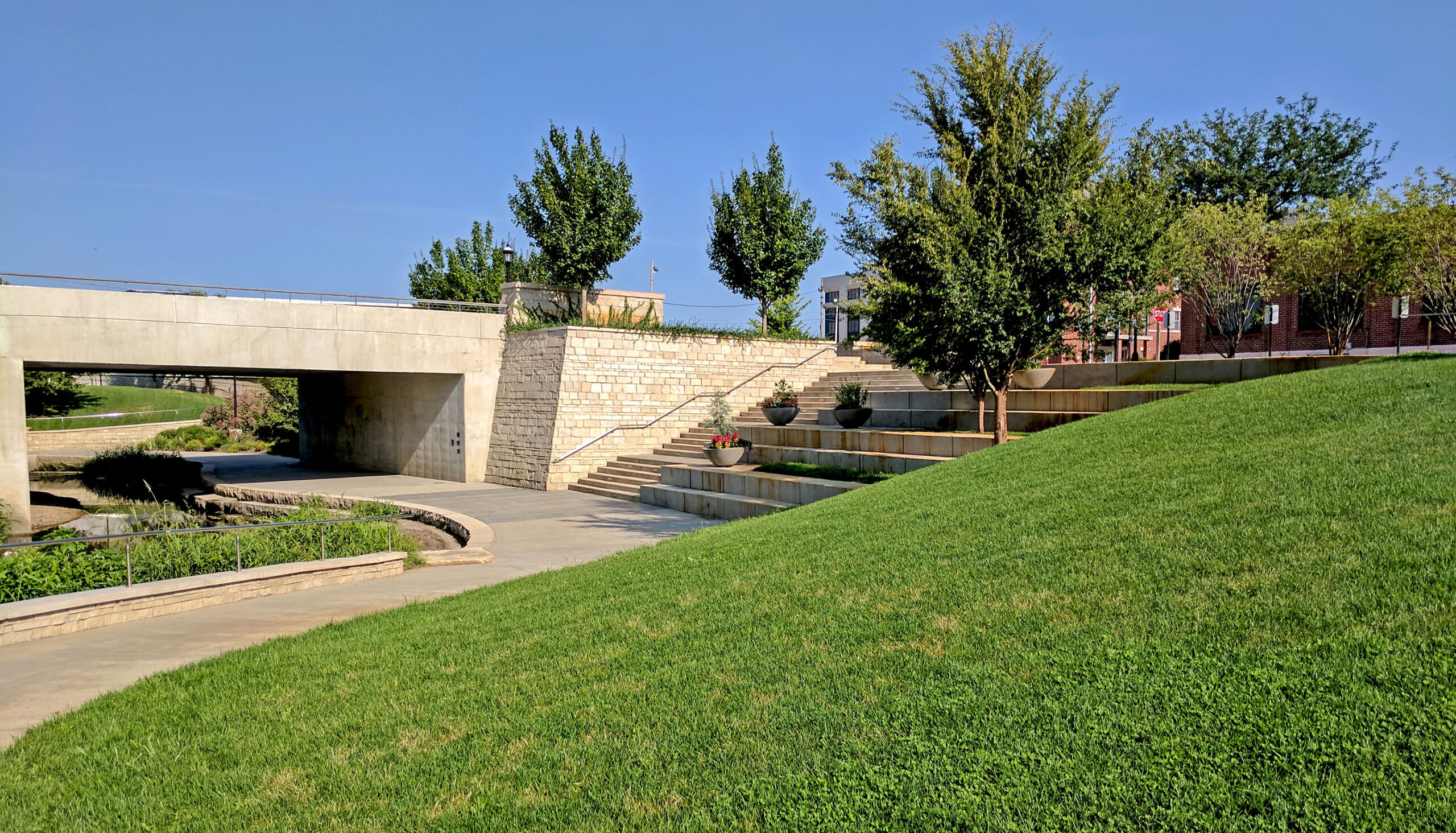 A green lawn slopes to the creek, terraces and steps lead down to the creek edge and from the street and shopping district above.