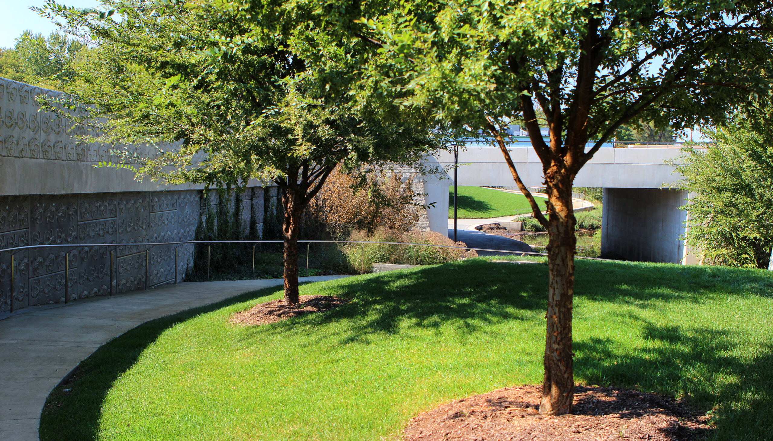 A path winds from the top of the urban area down to the creek edge and back up past stamped patterned walls and lush vegetation.