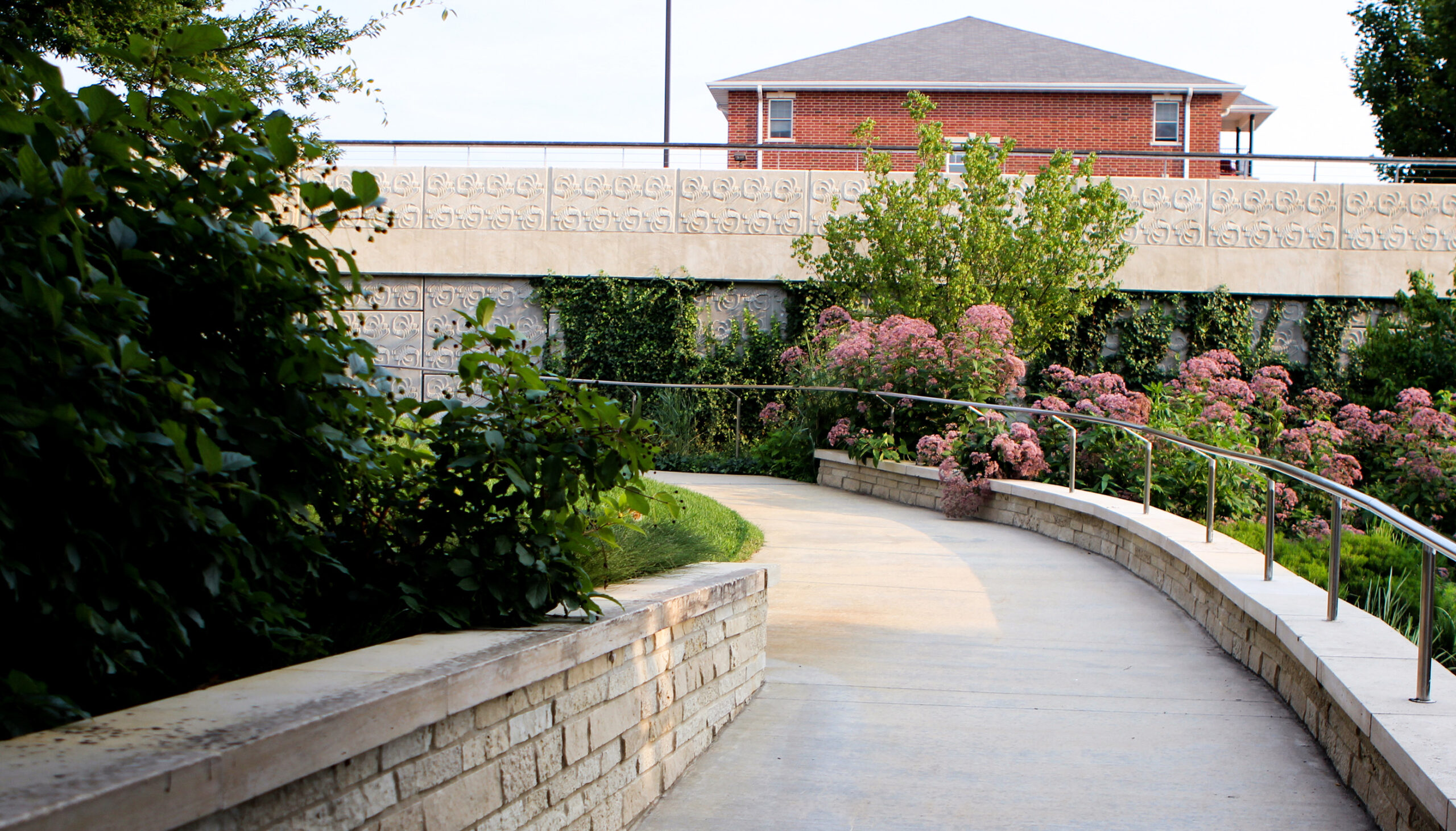 A path winds from the top of the urban area down to the creek edge and back up past stamped patterned walls and lush vegetation dotted with pink flowers.