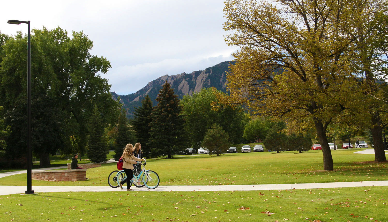 Two students walk their bikes by the turf green with the Flat Iron mountains in the background.
