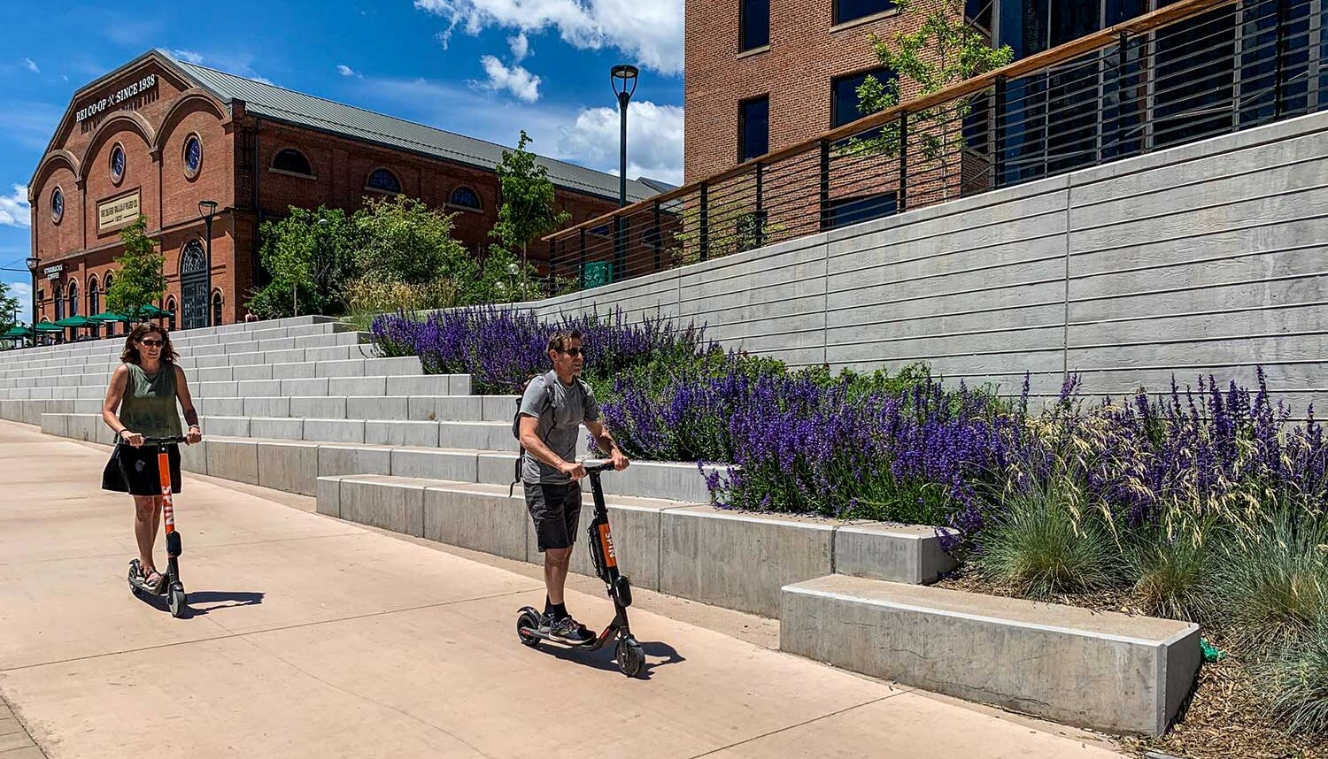 Two people scooter down the wide pedestrian walk between the terraced areas with purple flowers behind.