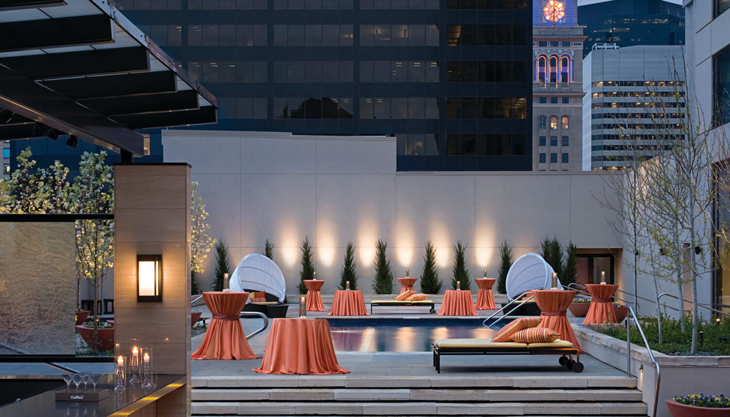 A photo of a rooftop pool terrace with juniper bushes against a uplit wall.
