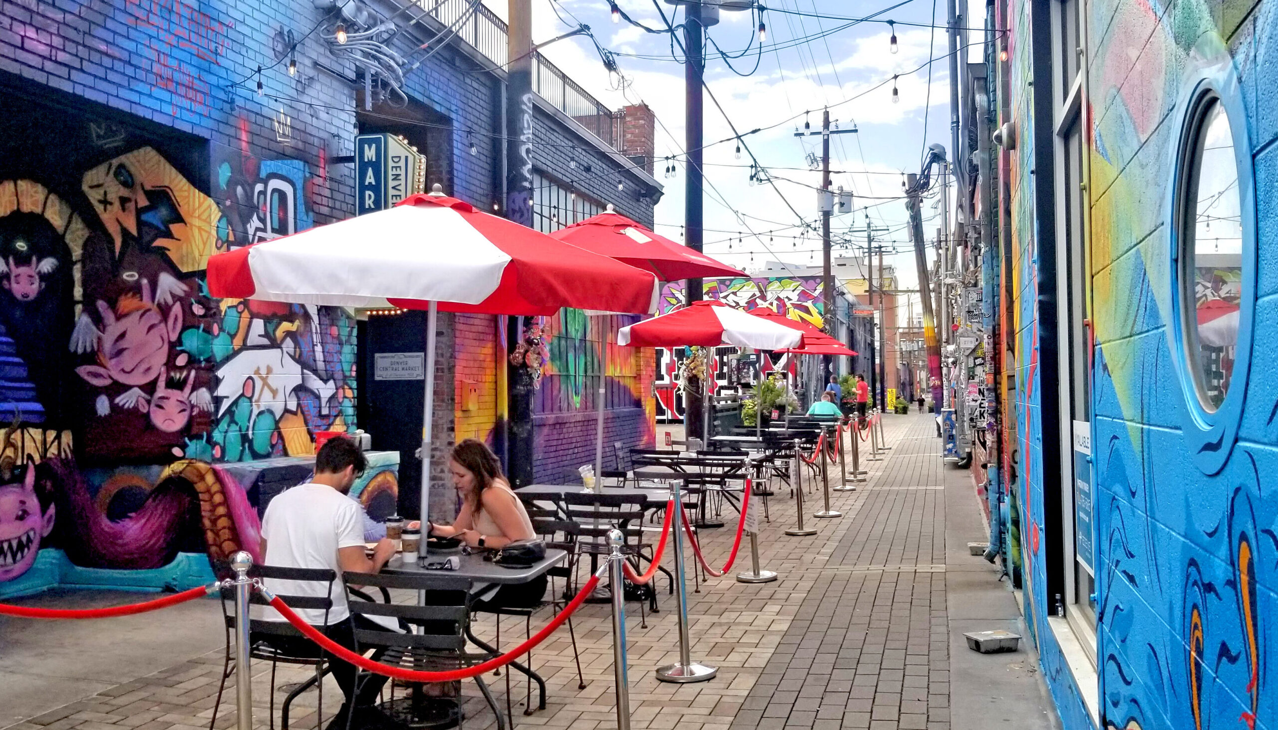 Mural-covered walls enclose a brick-paved alley with cafe seating and umbrellas.