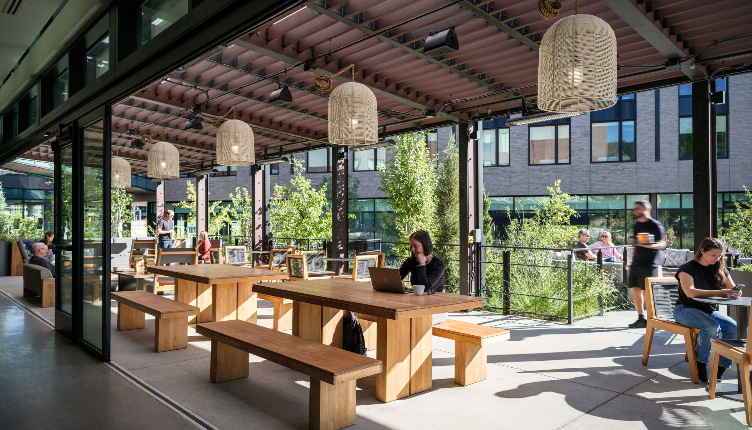 Indoor/outdoor community hotel patio with large wood tables under a canopy, decorative hanging lamps, and plantings.