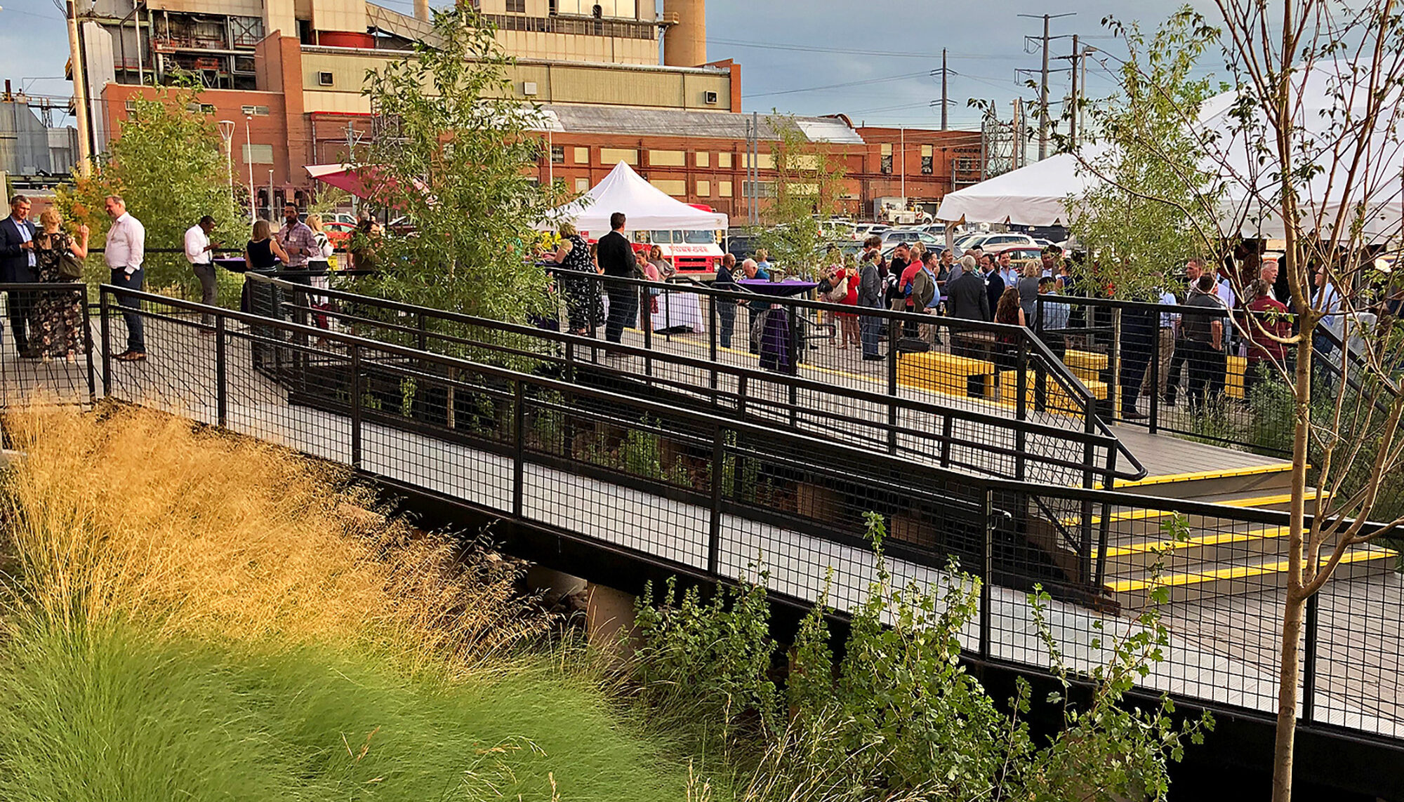 a party on the plaza in the background, in the foreground pedestrian bridges cross over a sunken rain garden filled with native grasses and young cottonwood trees