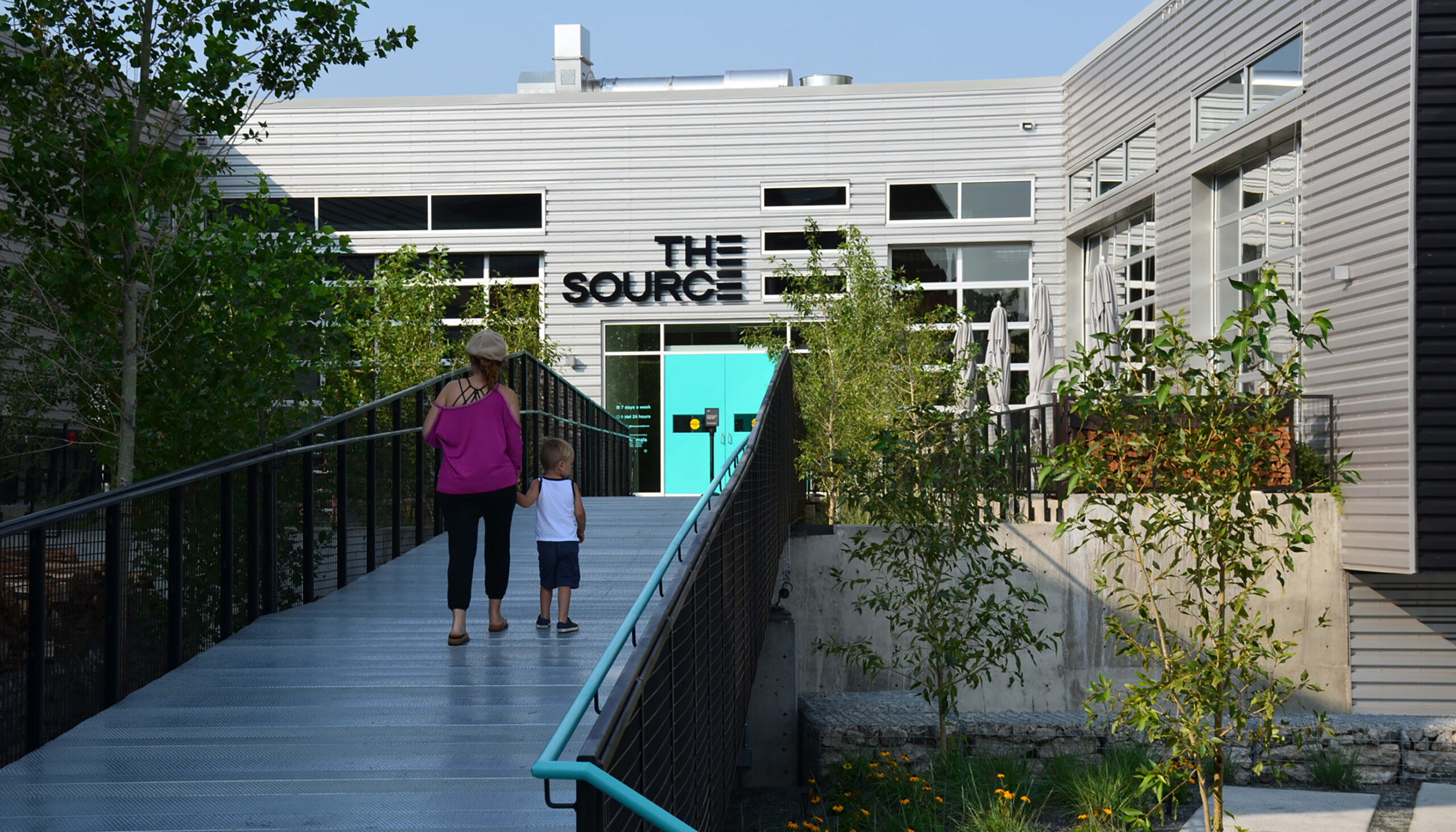 A pedestrian bridge leads between the tops of trees to the turquoise door of the Source Hotel