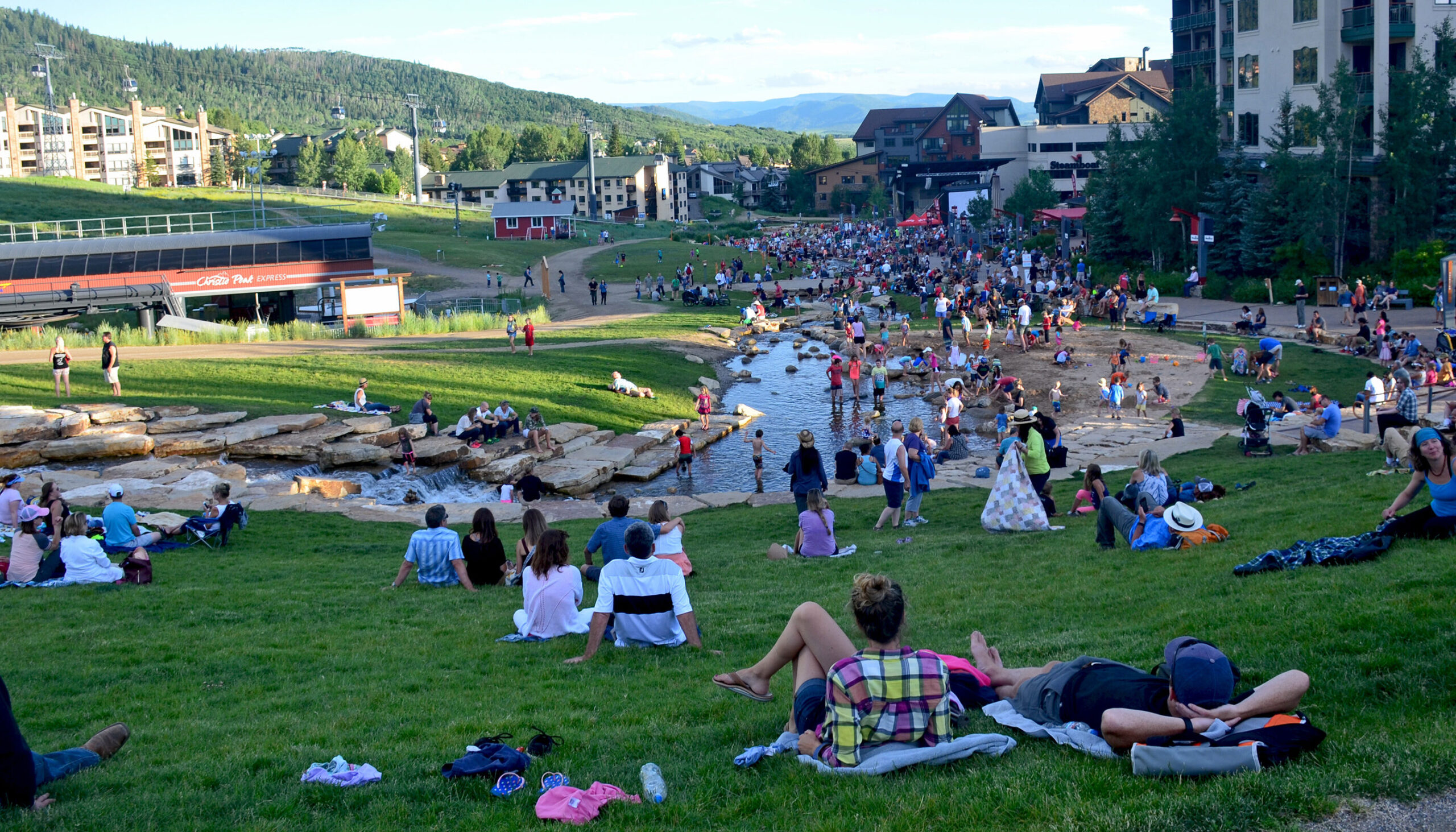 view from the green grass slope looking down on the crowded Burgess Creek beach area and the mountain village and mountains beyond on a warm summer evening
