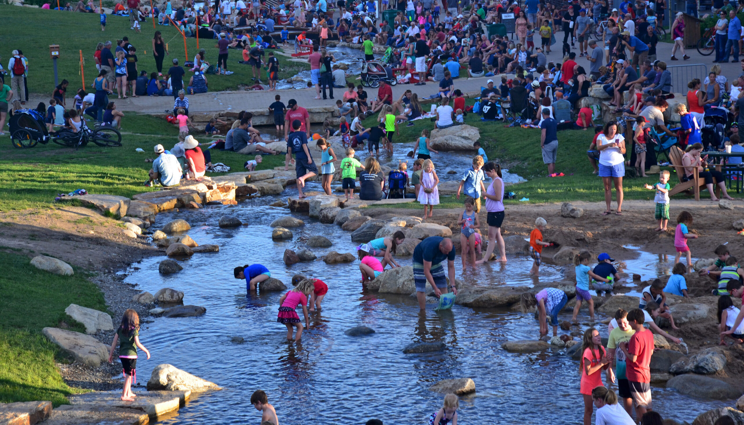 large crowd on a summer night fills the edges of the creek and promenade, people wade in the water at the beach area in the foreground