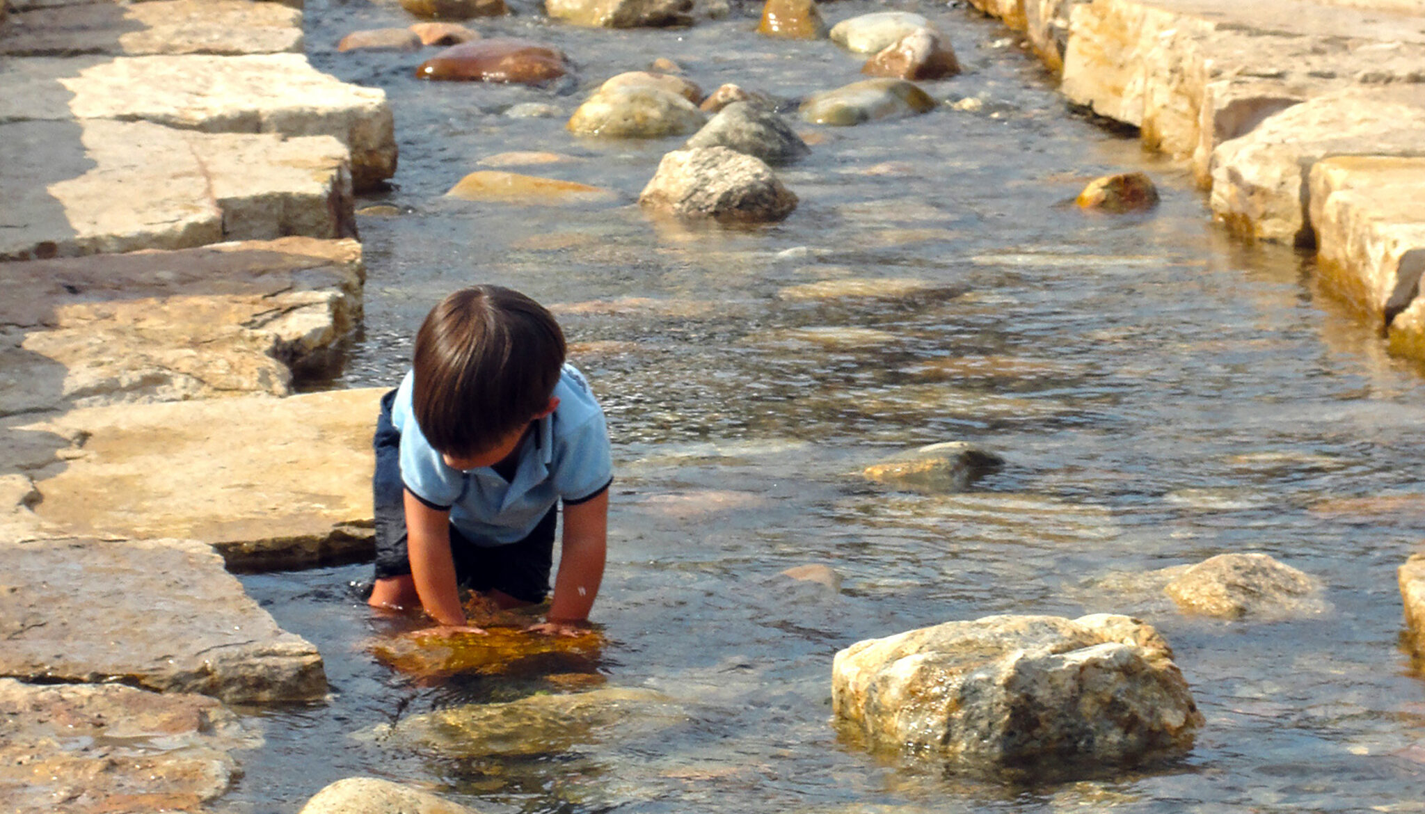 a child in a blue shirt plays in the shallow water next to the stone edge of Burgess Creek