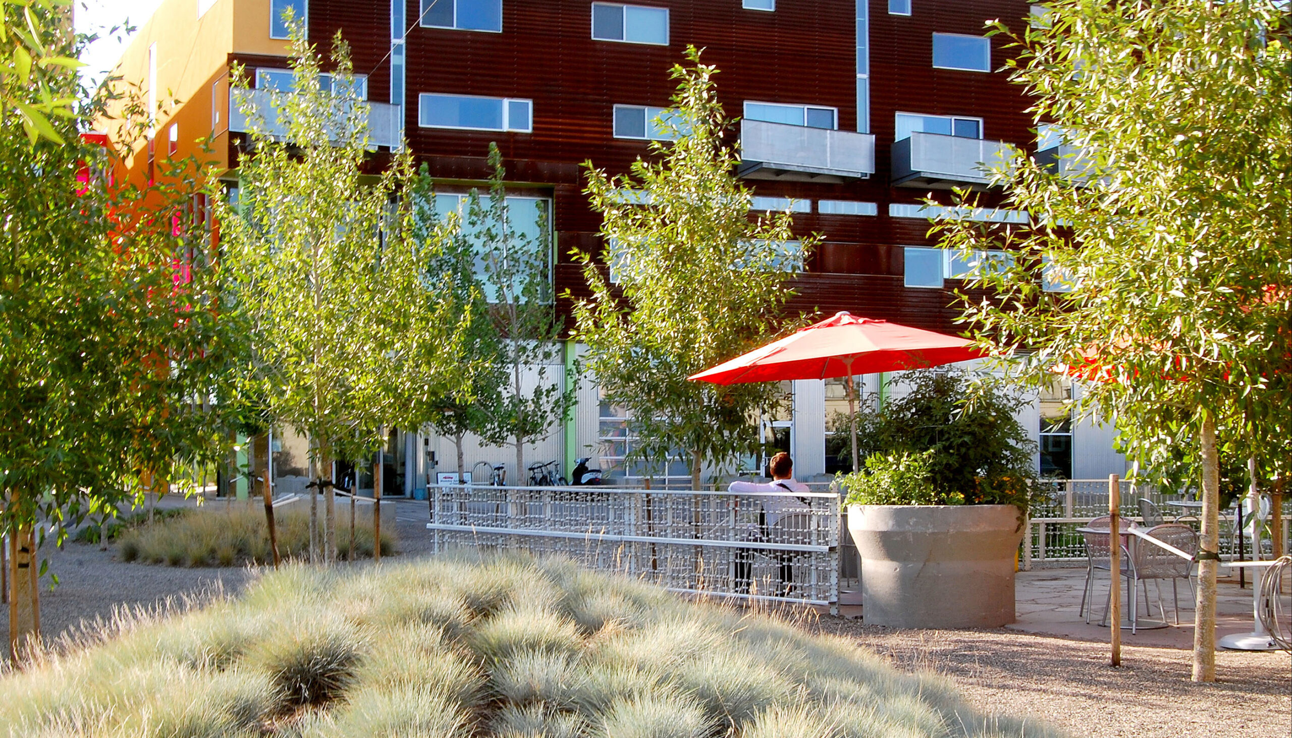 person sitting under a red umbrella at a cafe within the mixed-used development surrounded by poplar trees and mounds of blue fescue grasses