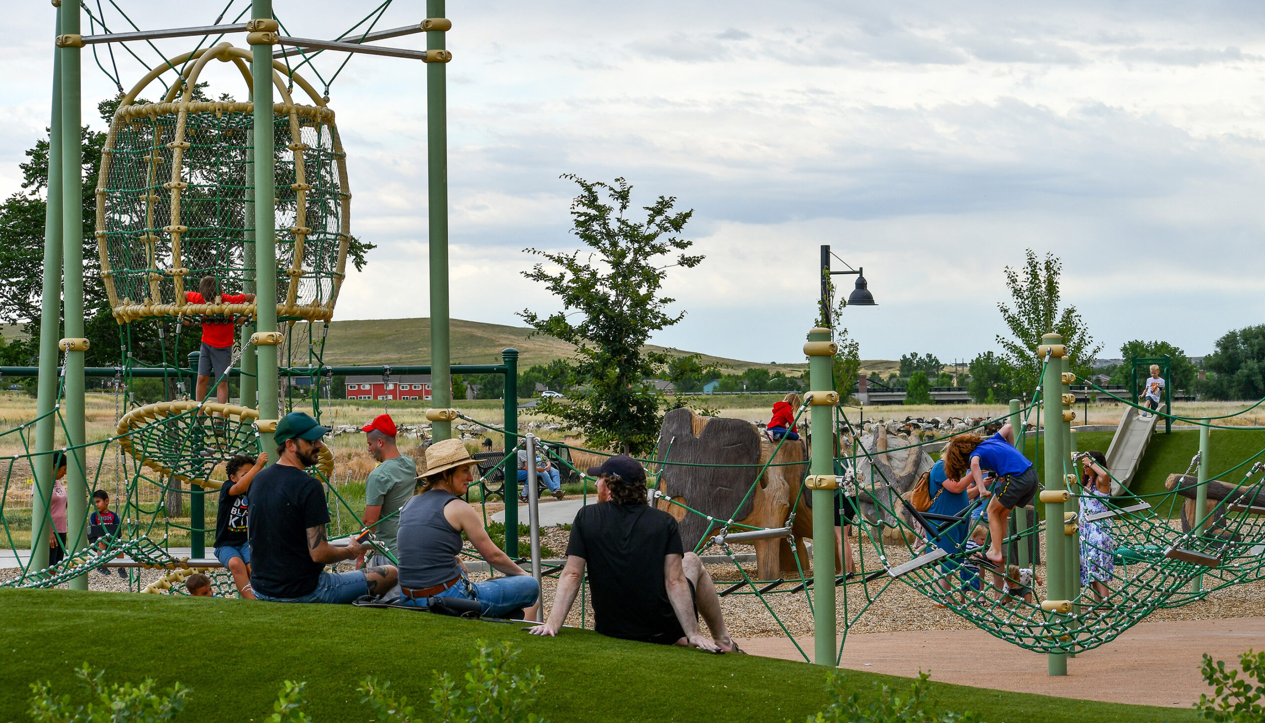 parents sit on a turf slope watching their kids on the rope play equipment