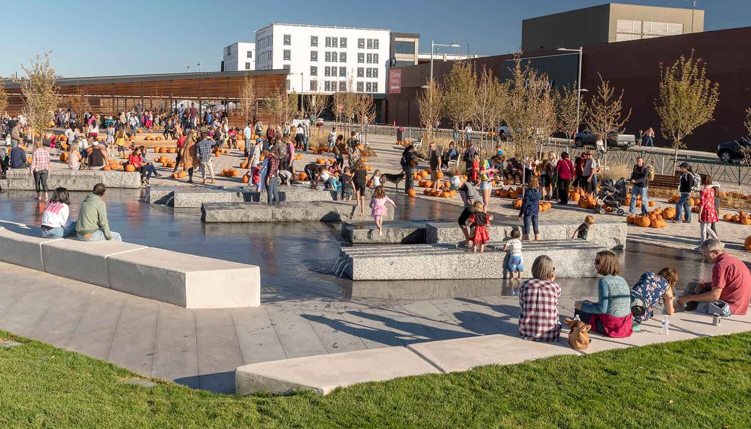 A community fall festival takes place on the central plaza, with pumpkins scattered all over, and kids splashing through the granite water feature while their parents look on.